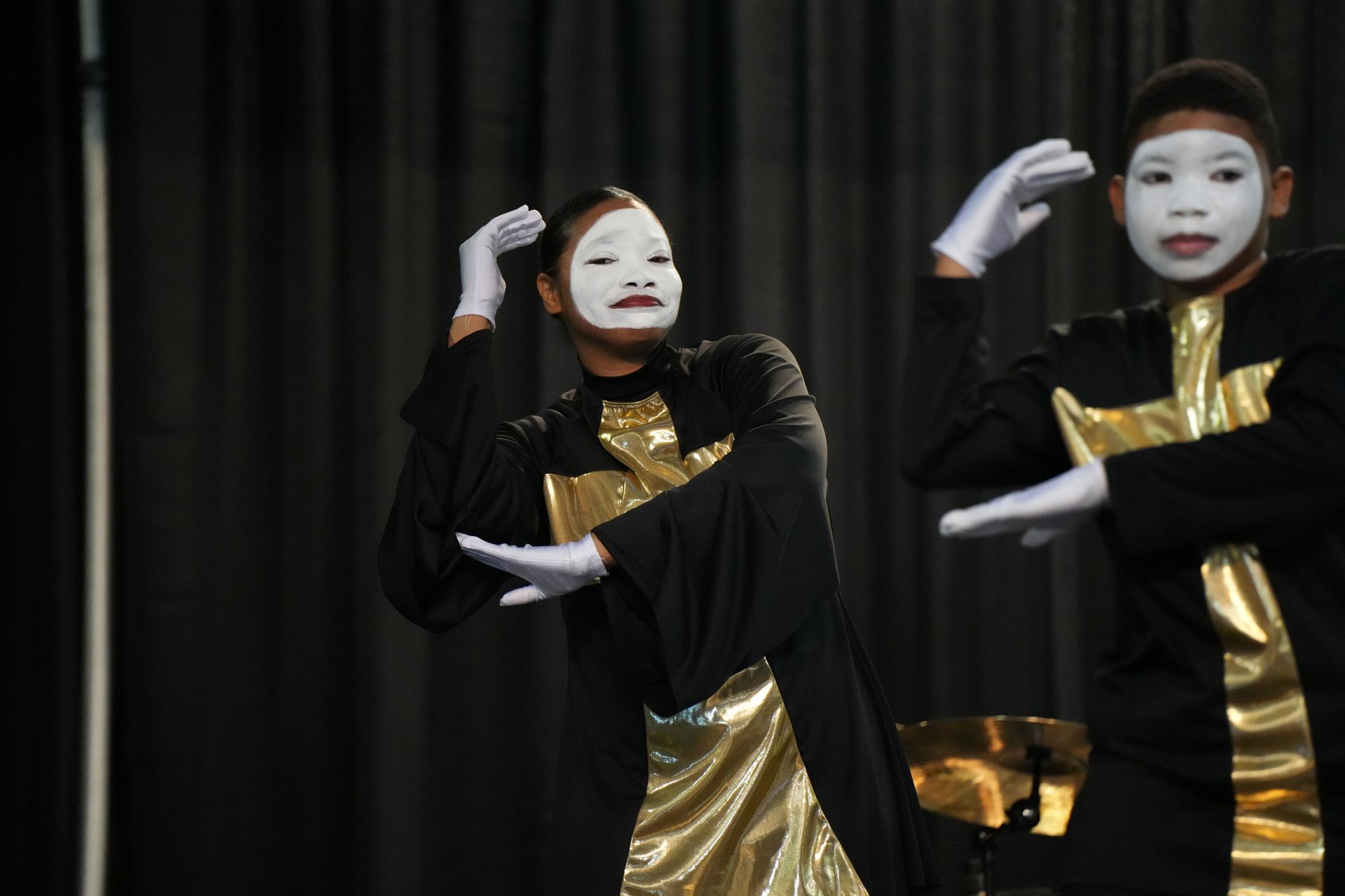 Two dancers in white masks and black-and-gold costumes perform on a dark stage