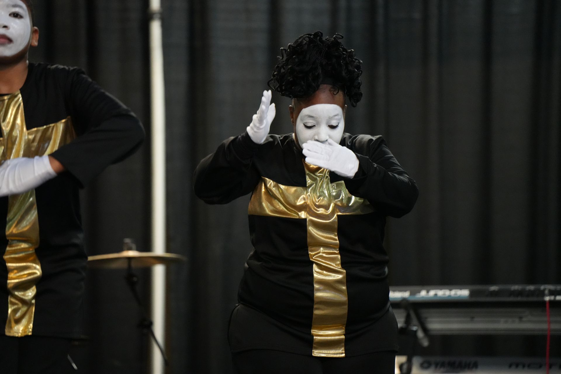 Performer in black and gold costume with white face paint, posed on stage with another performer partially visible.