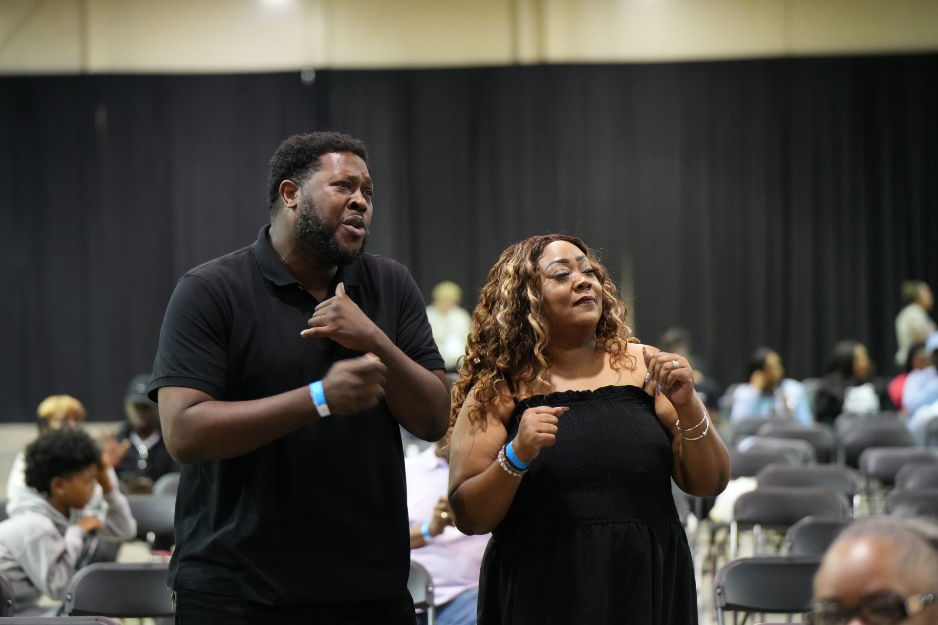 Two people in black outfits standing and speaking in a conference hall with seated attendees