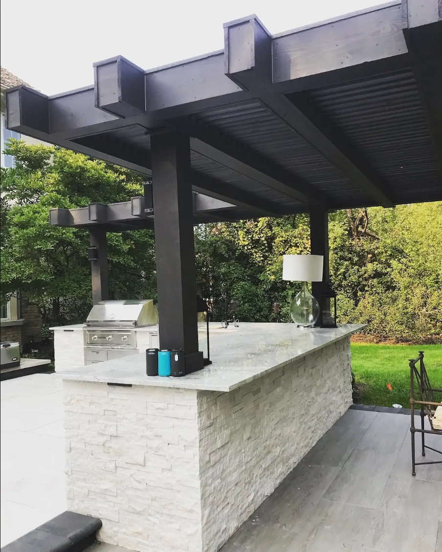 Outdoor kitchen with dark pergola, white stone base, and marble countertop. Grill and bar seating.