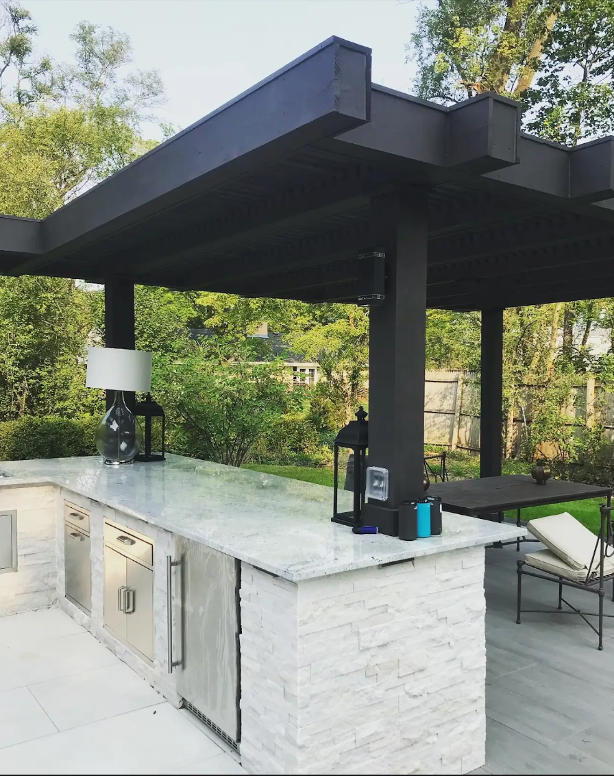 Outdoor kitchen with black pergola, white brick, stainless steel appliances, and marble countertop.