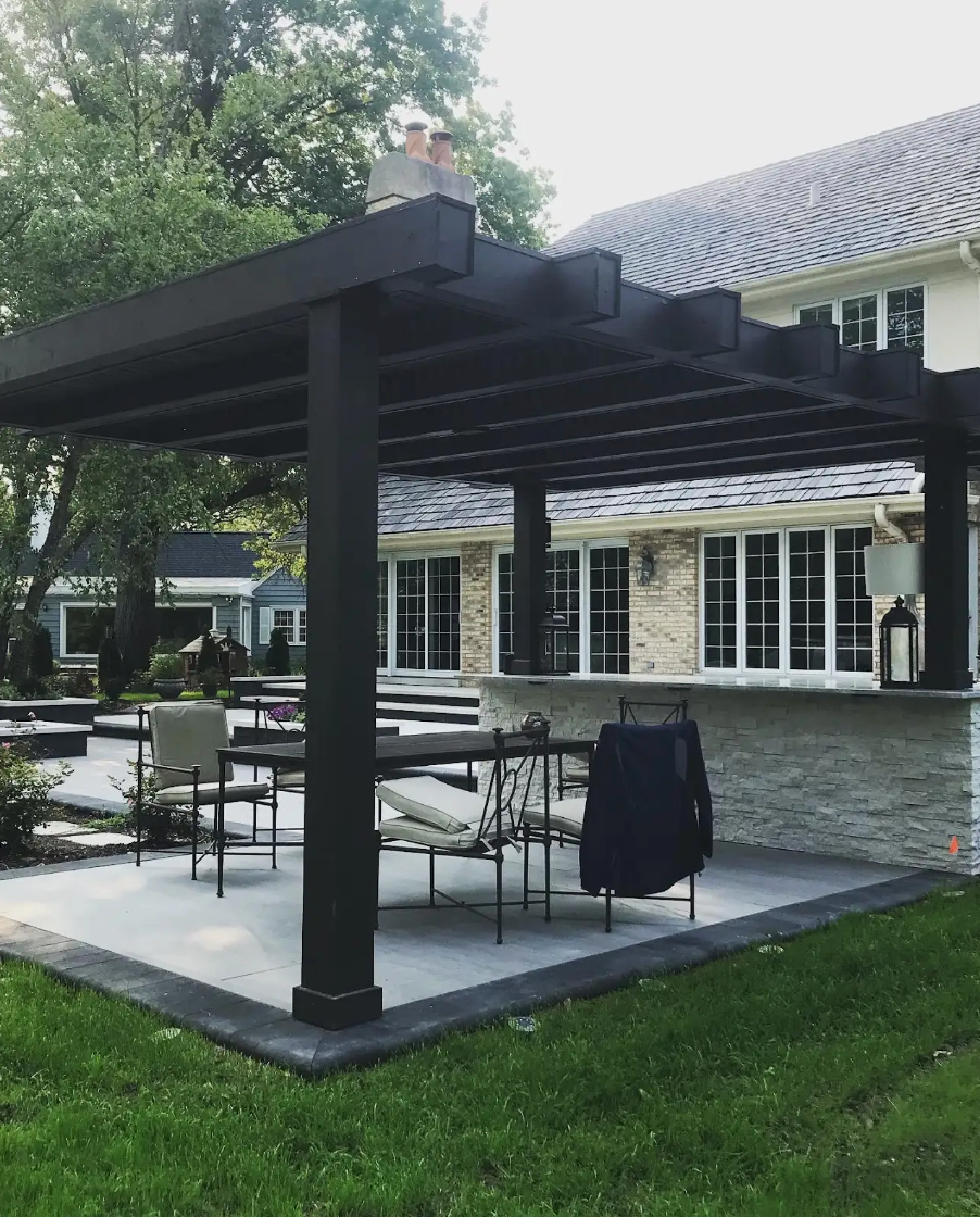 Black pergola over a patio with a bar and seating, next to a brick house.