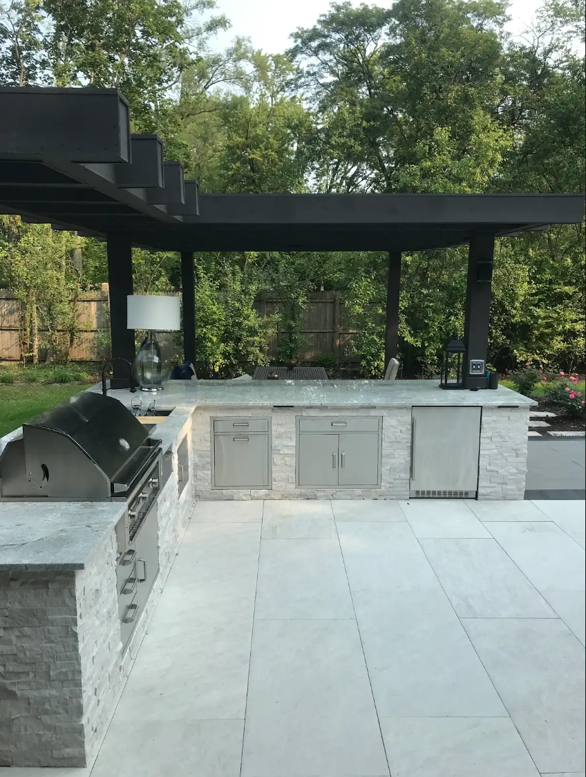 Outdoor kitchen with grill, countertops, and overhead pergola, surrounded by trees.