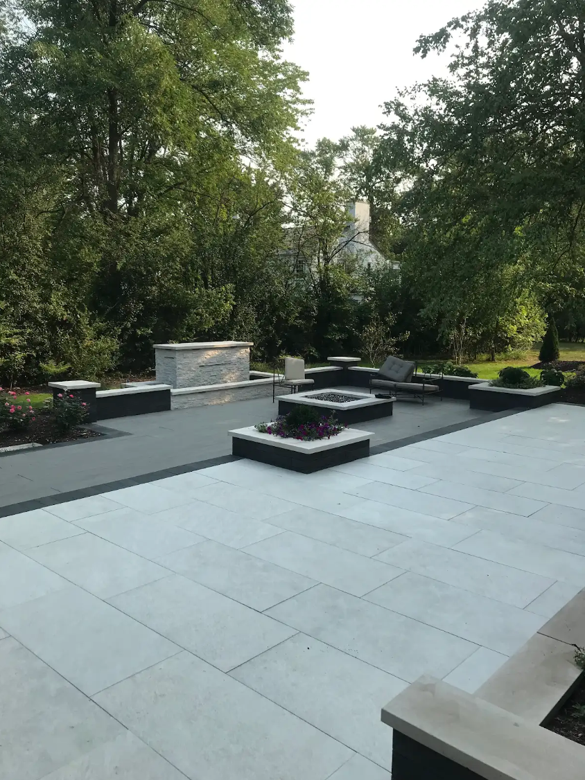 Patio with gray stone pavers, built-in seating, fire pits, and a built-in kitchen area, surrounded by trees.
