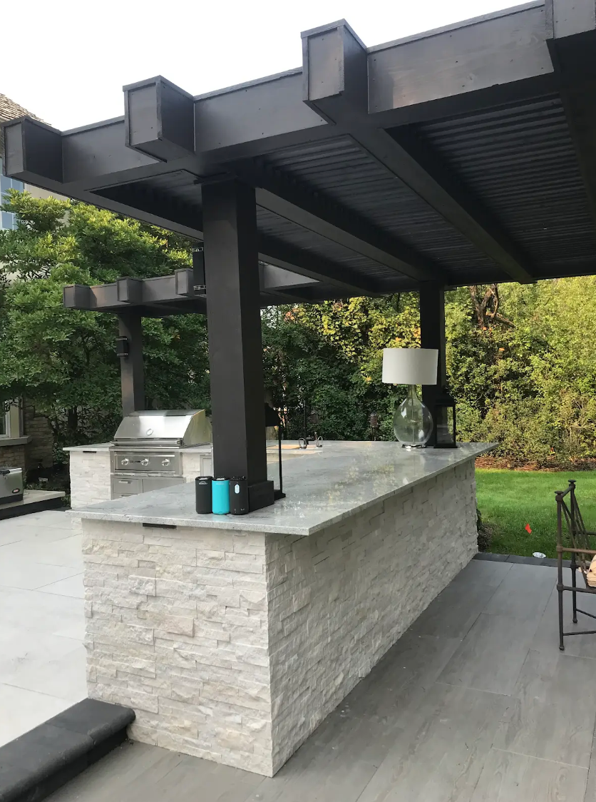 Outdoor kitchen with stone-faced island, granite countertop, pergola, and grill.