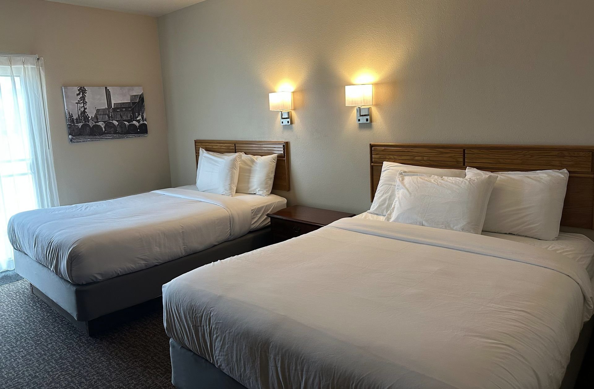 Two beds in a hotel room with white bedding, a nightstand, and wall sconces.