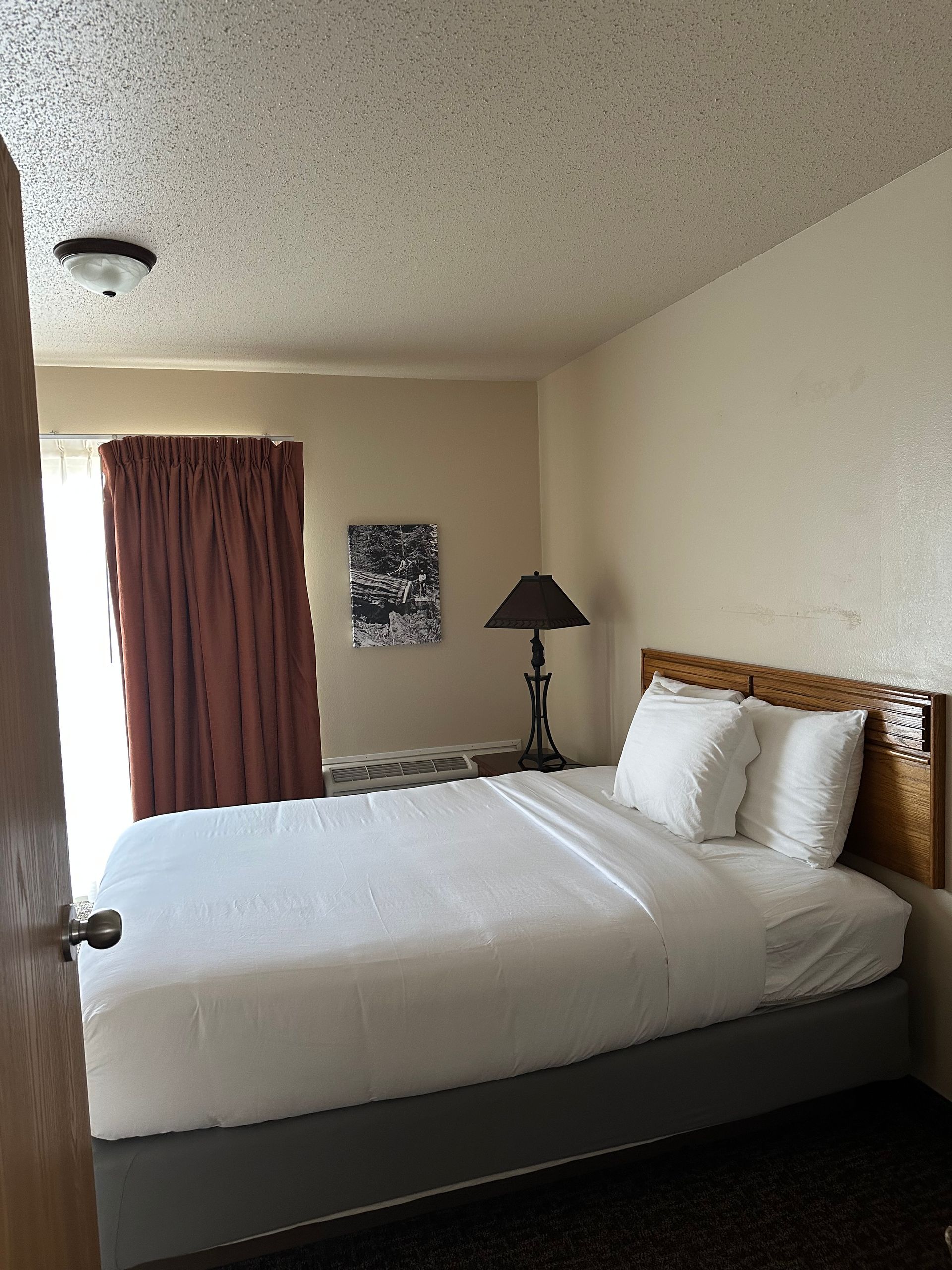 A hotel room with a bed, lamp, and curtains. Brown, white, beige tones.