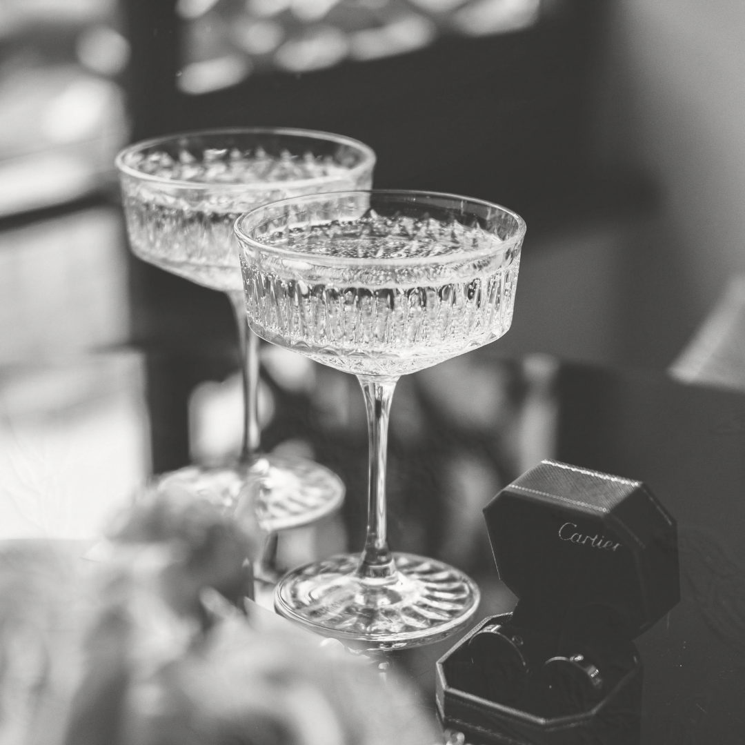 A black and white photo of two champagne glasses and an engagement ring