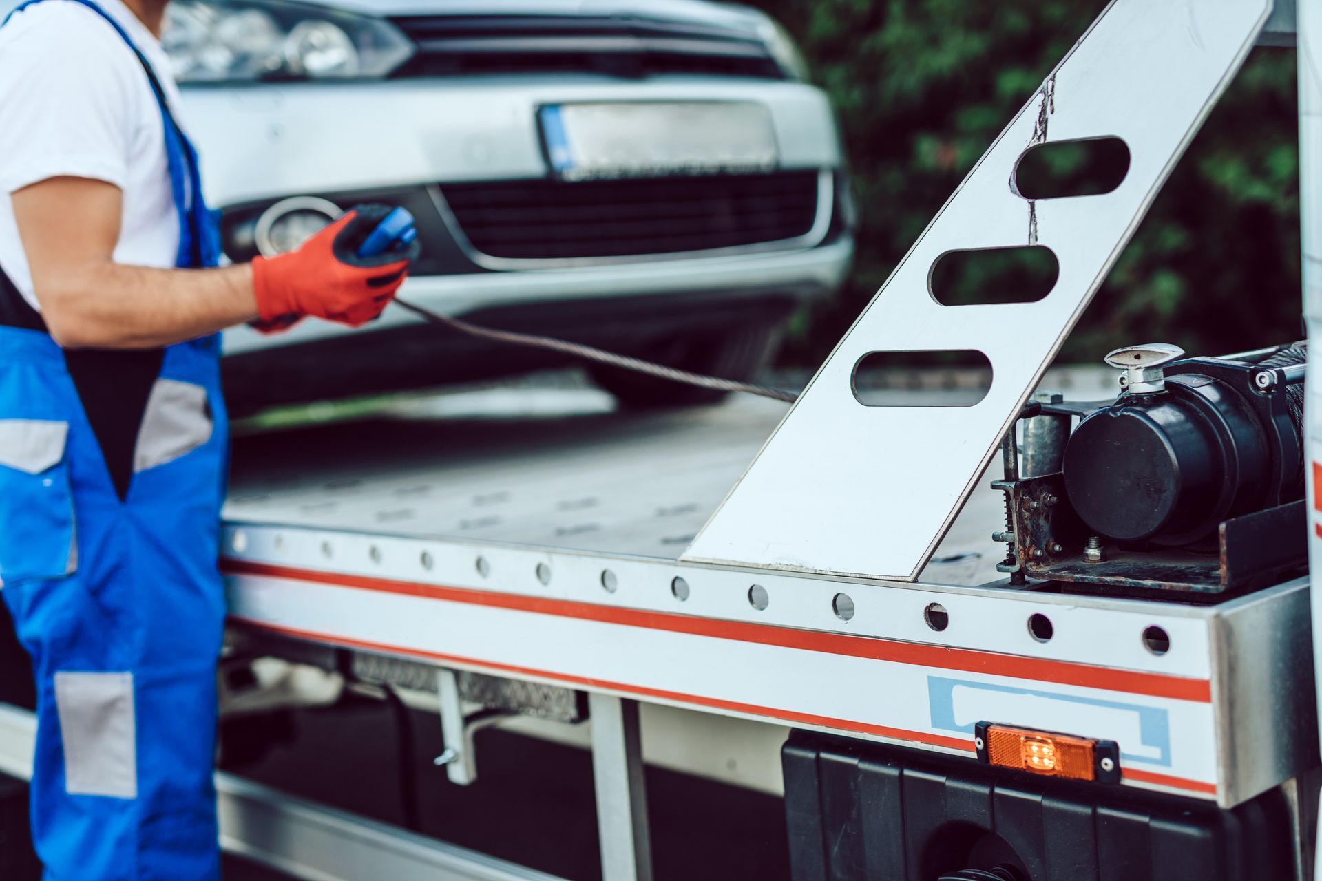 Tow truck operator in blue overalls attaching a car to a flatbed tow truck