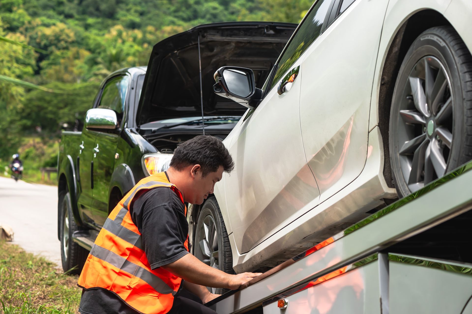 Tow truck operator in orange vest loading a white car onto a flatbed on a roadside