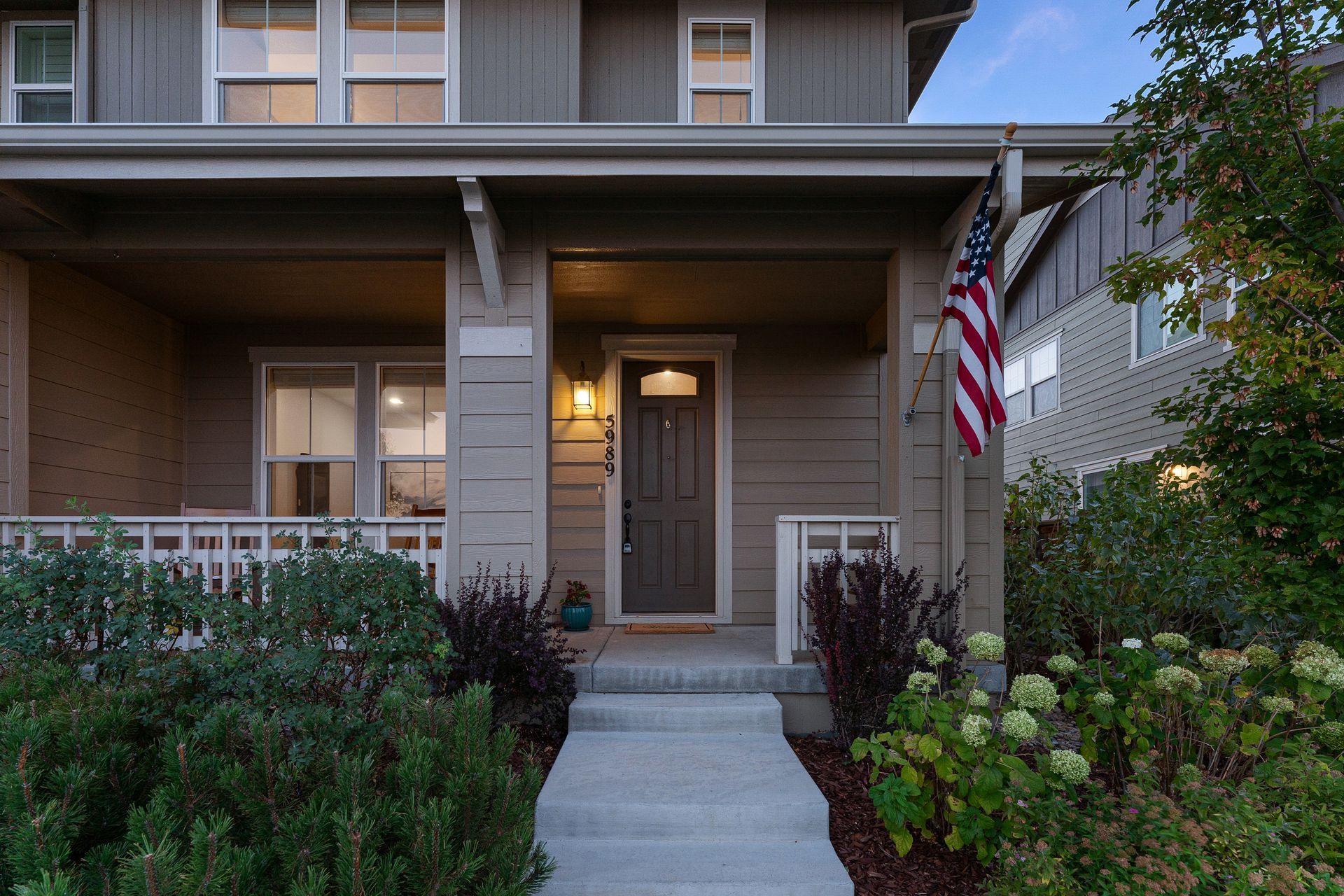 House with a front porch, American flag, and landscaping. Gray siding, front door, and steps.