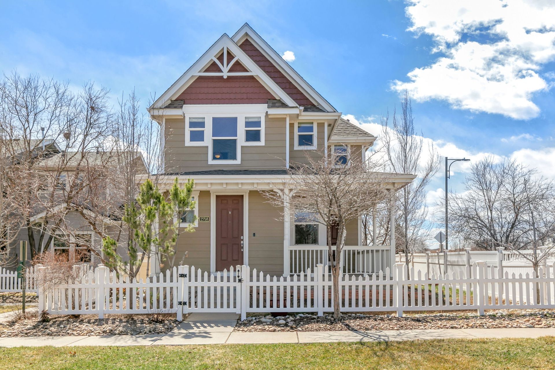 Tan two-story house with a white picket fence under a blue sky with trees.
