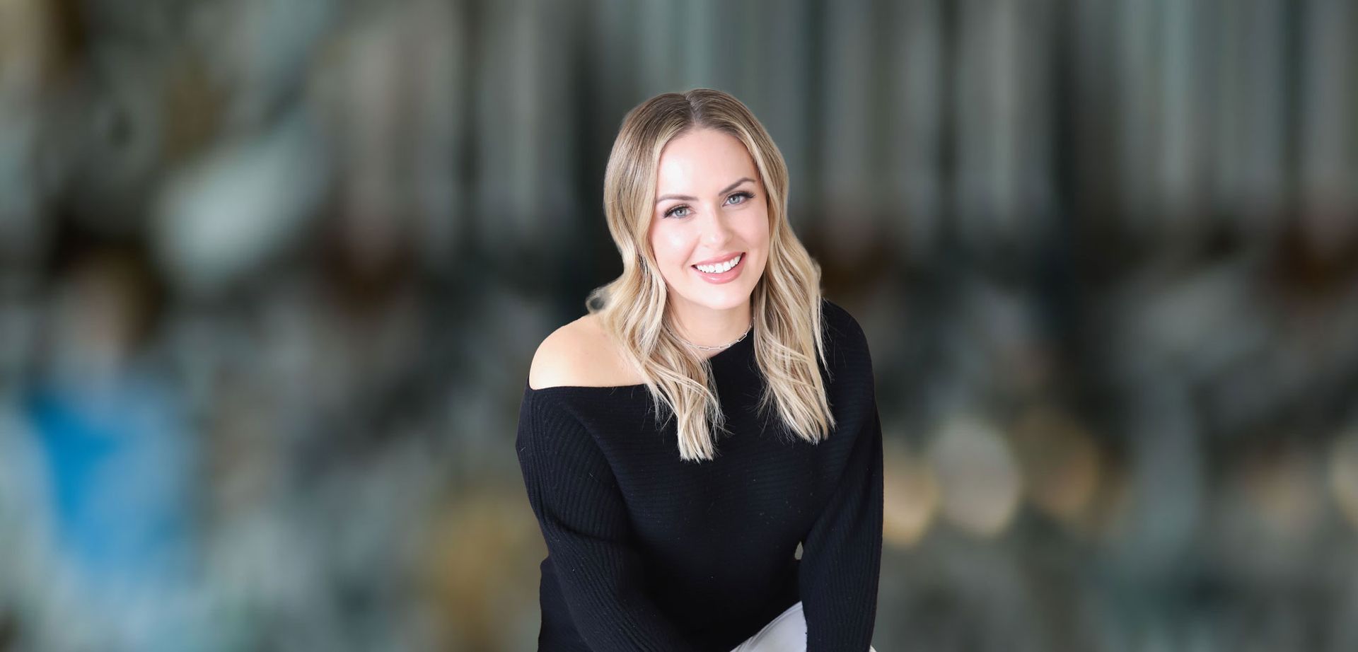 Woman with blonde hair wearing a black off-the-shoulder sweater smiles at the camera against a blurred background.