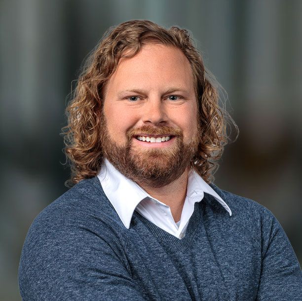 Man with curly blond hair, beard, wearing blue sweater over white collared shirt, smiling, business-like.