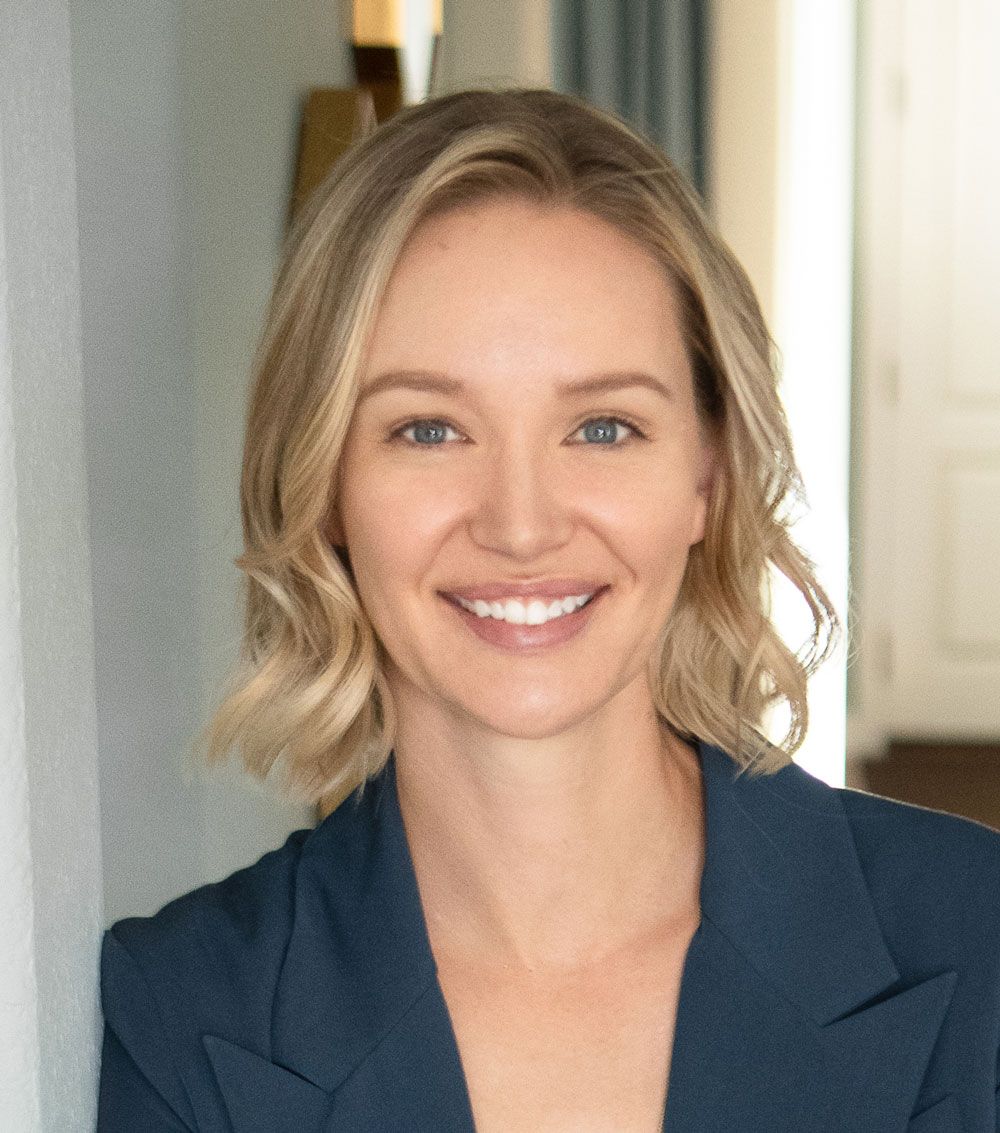 A woman in a blue jacket is smiling and leaning against a wall.