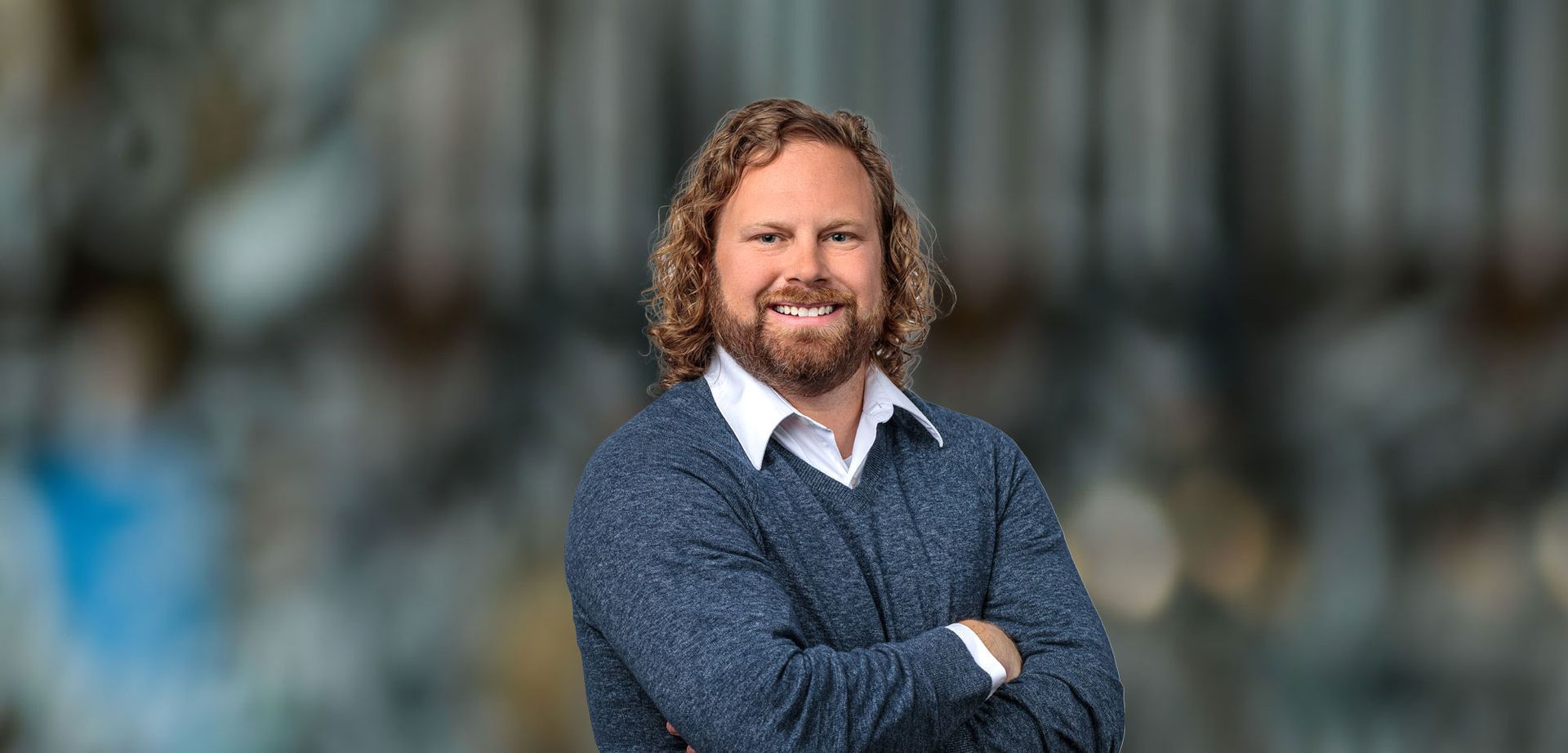 Man with curly hair and beard smiles, arms crossed, wearing blue sweater over white shirt, against blurred background.