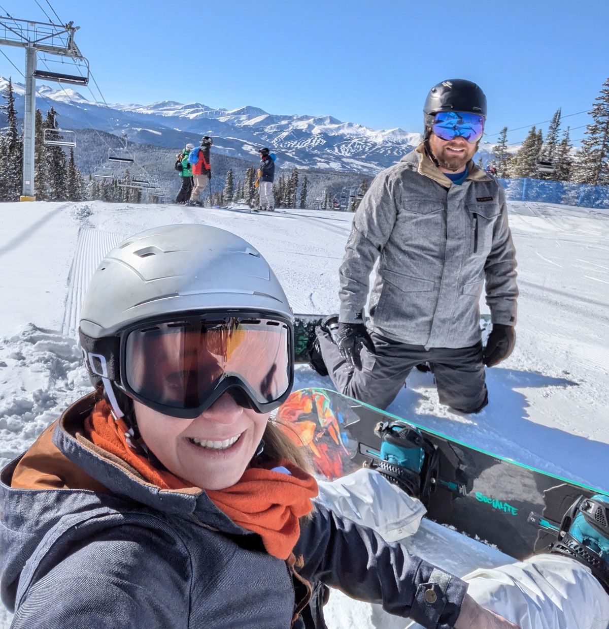 Two people on a snowy slope with snowboard. One kneels, the other smiles with goggles and a helmet. Mountains and ski lift in background.