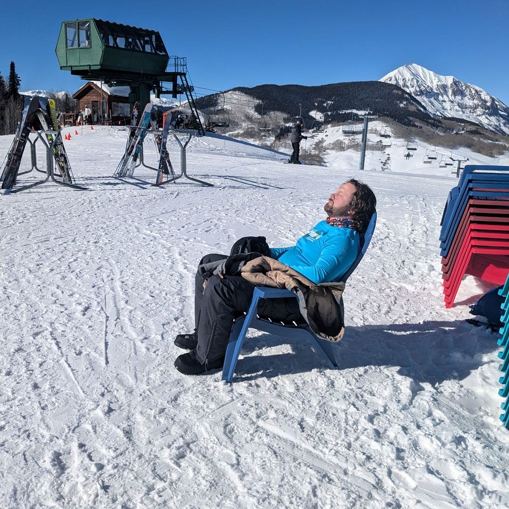Person in blue shirt relaxes in a chair on a snowy mountain. Ski lift and mountain in the background.