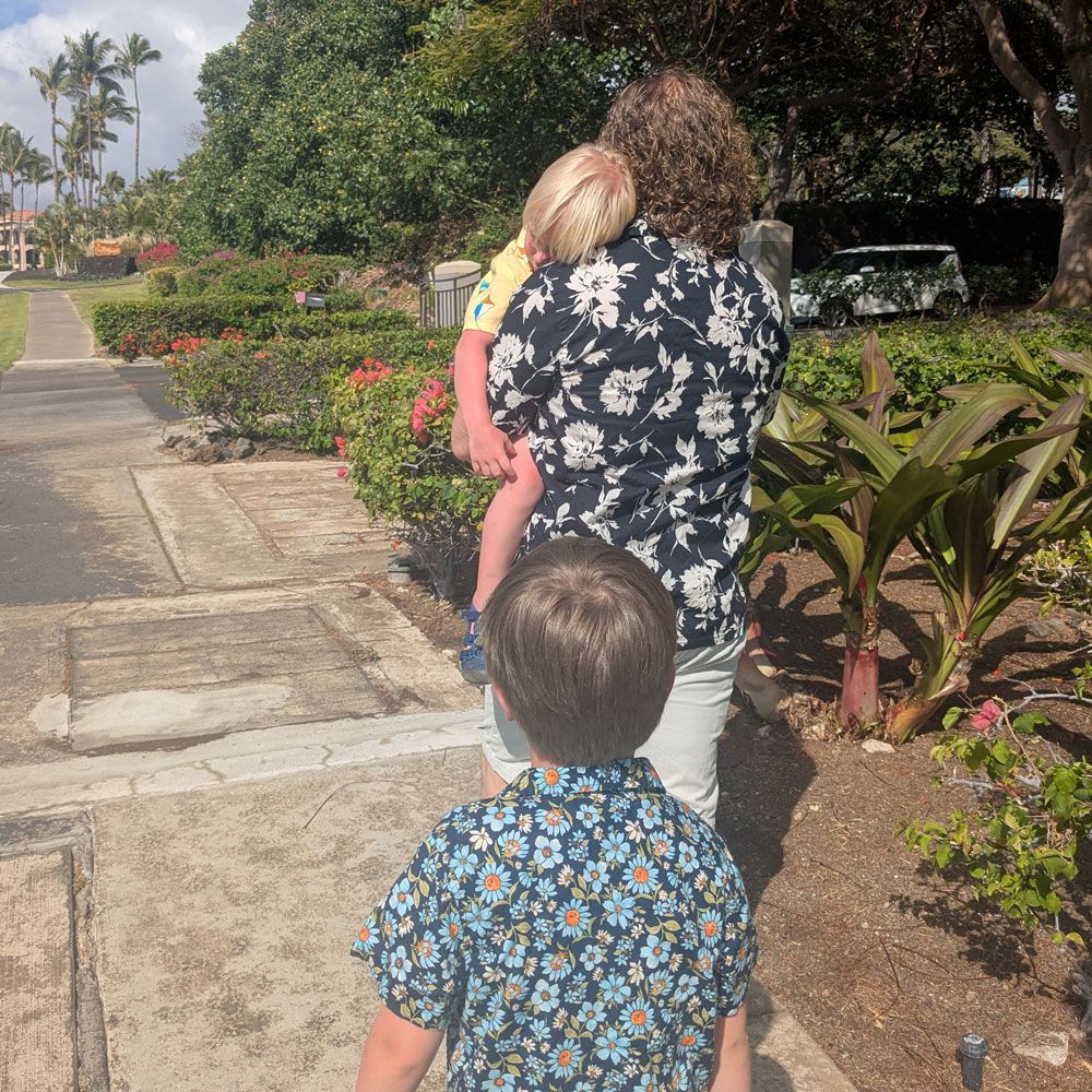 A person carrying a child walks down a sidewalk. Another child follows. They are near flowers and greenery.