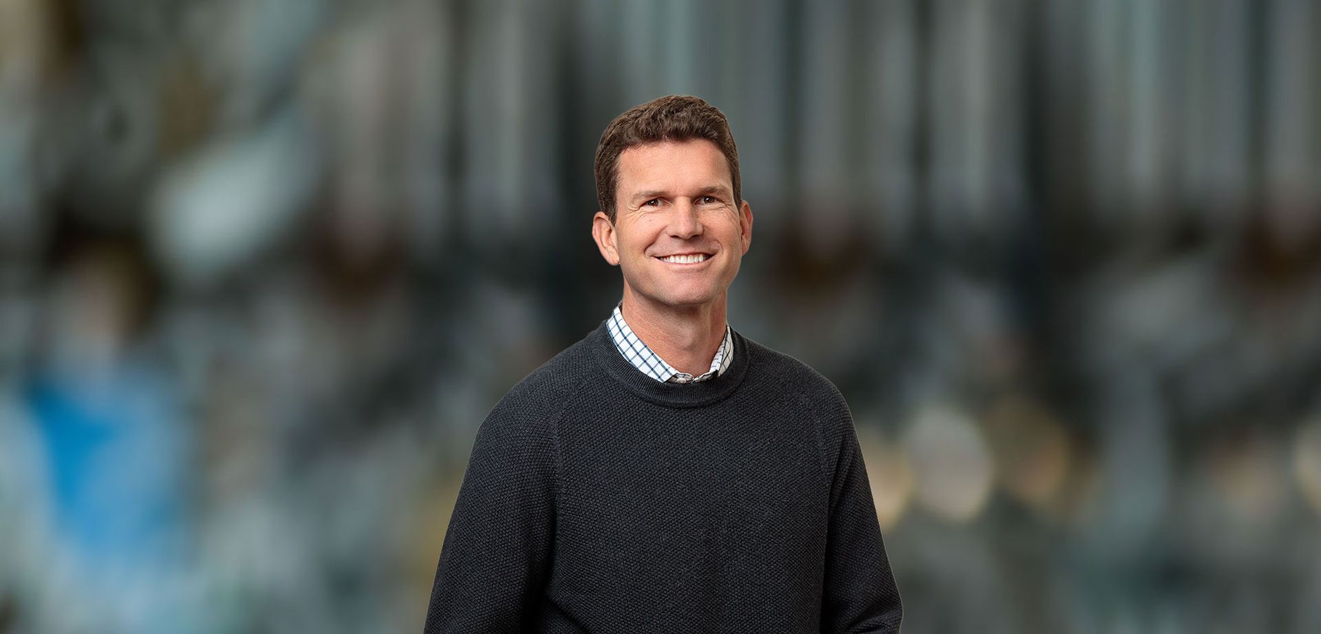 Man smiling, wearing a dark sweater and collared shirt, blurred background.