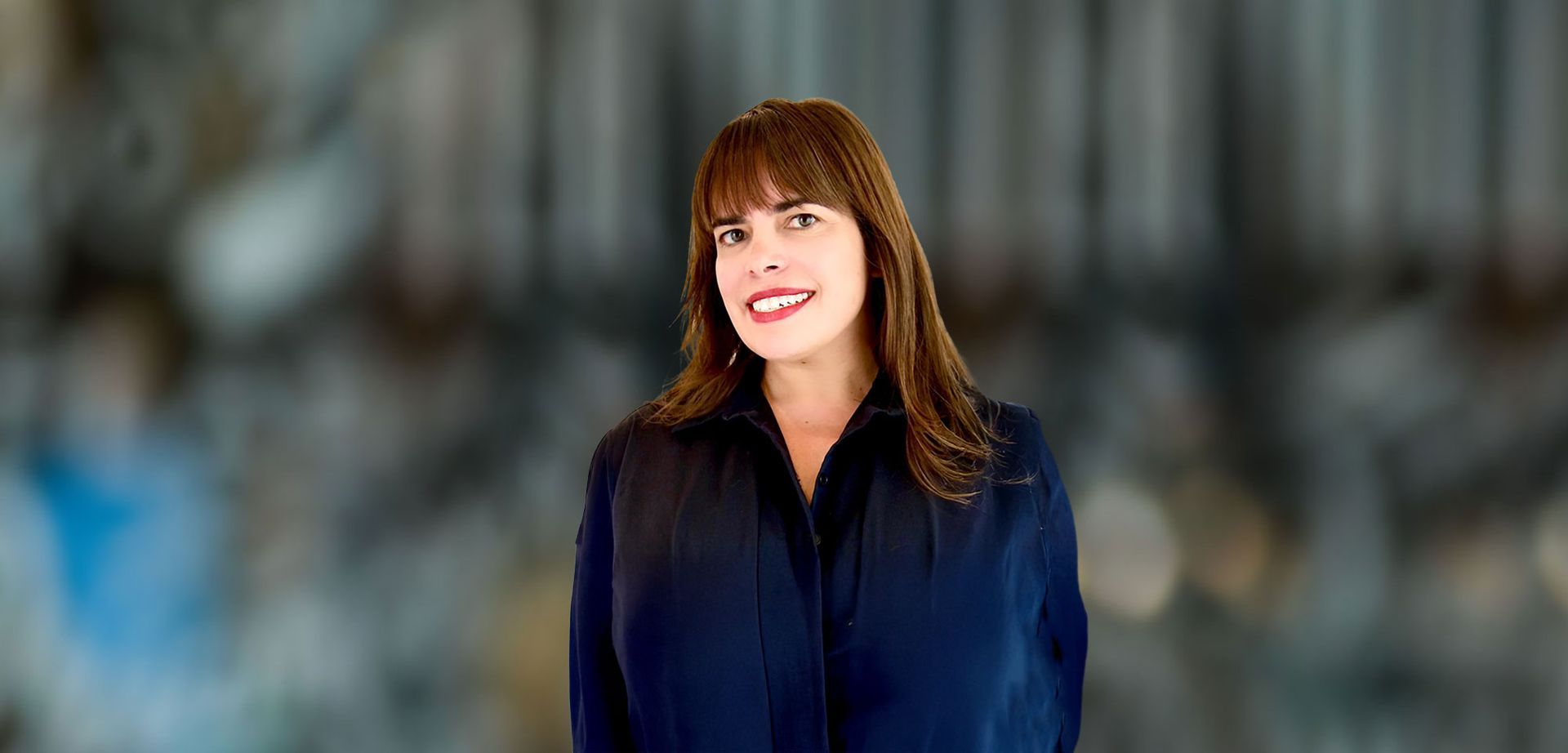 Woman with brown hair and bangs smiles, wearing a dark blue jacket, against a blurred background.