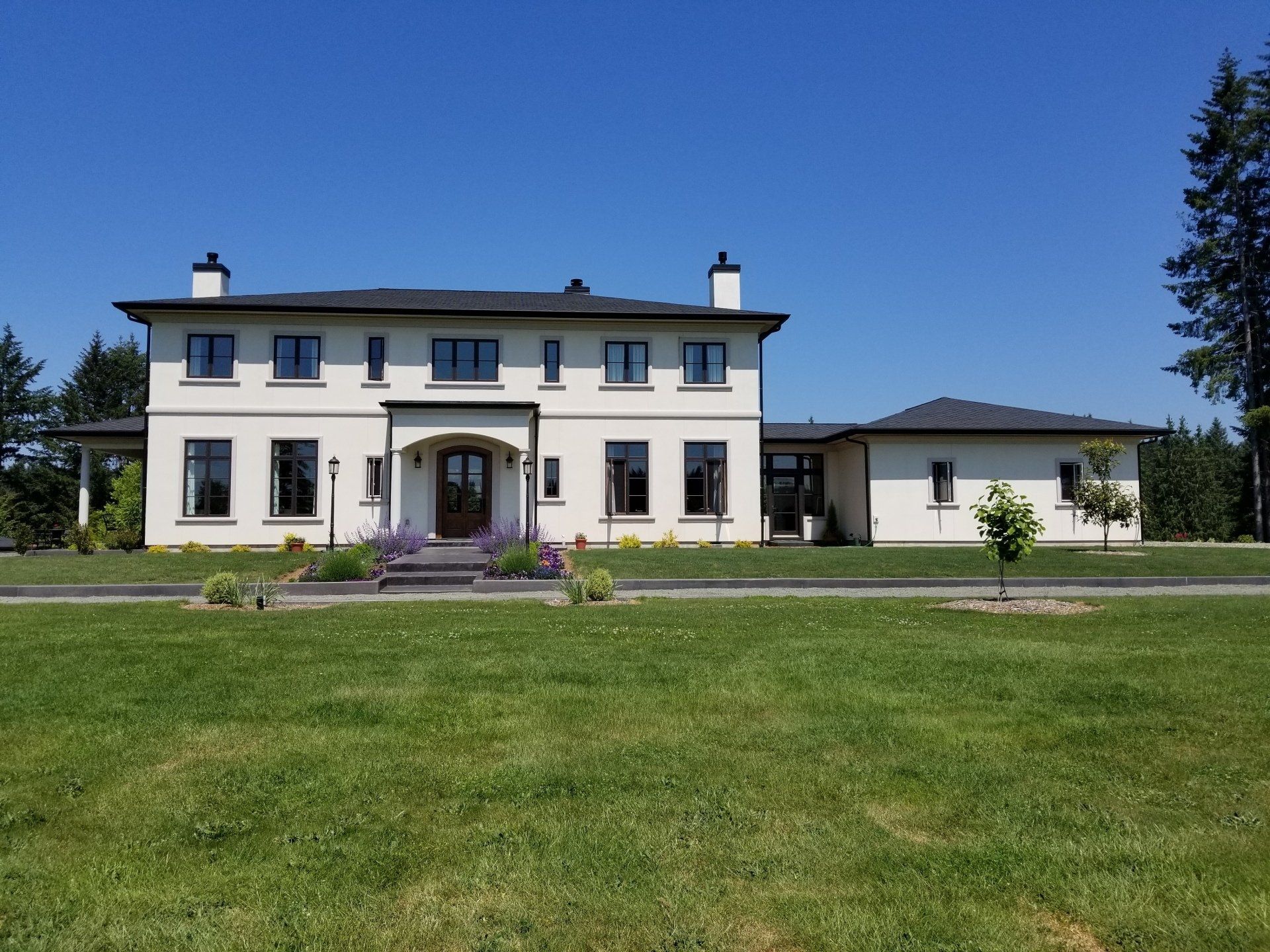 Two-story beige house with black-framed windows and doors, on a large grassy lawn under a blue sky.