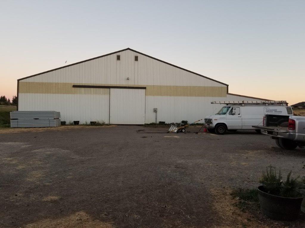 White barn with roll-up door, work van, and metal beams on a gravel lot at dusk.