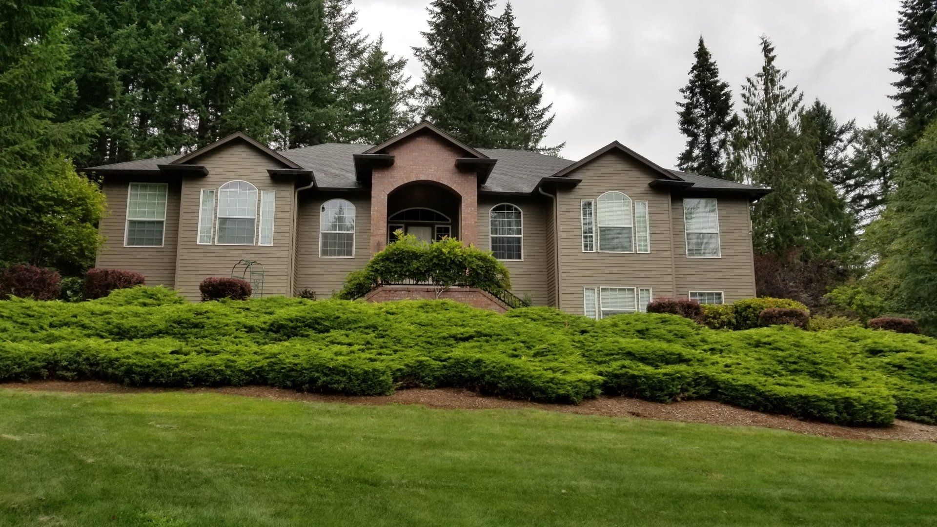 Two-story house with brick facade, arched entryway, and green shrubbery in front. Overcast sky and trees in the background.