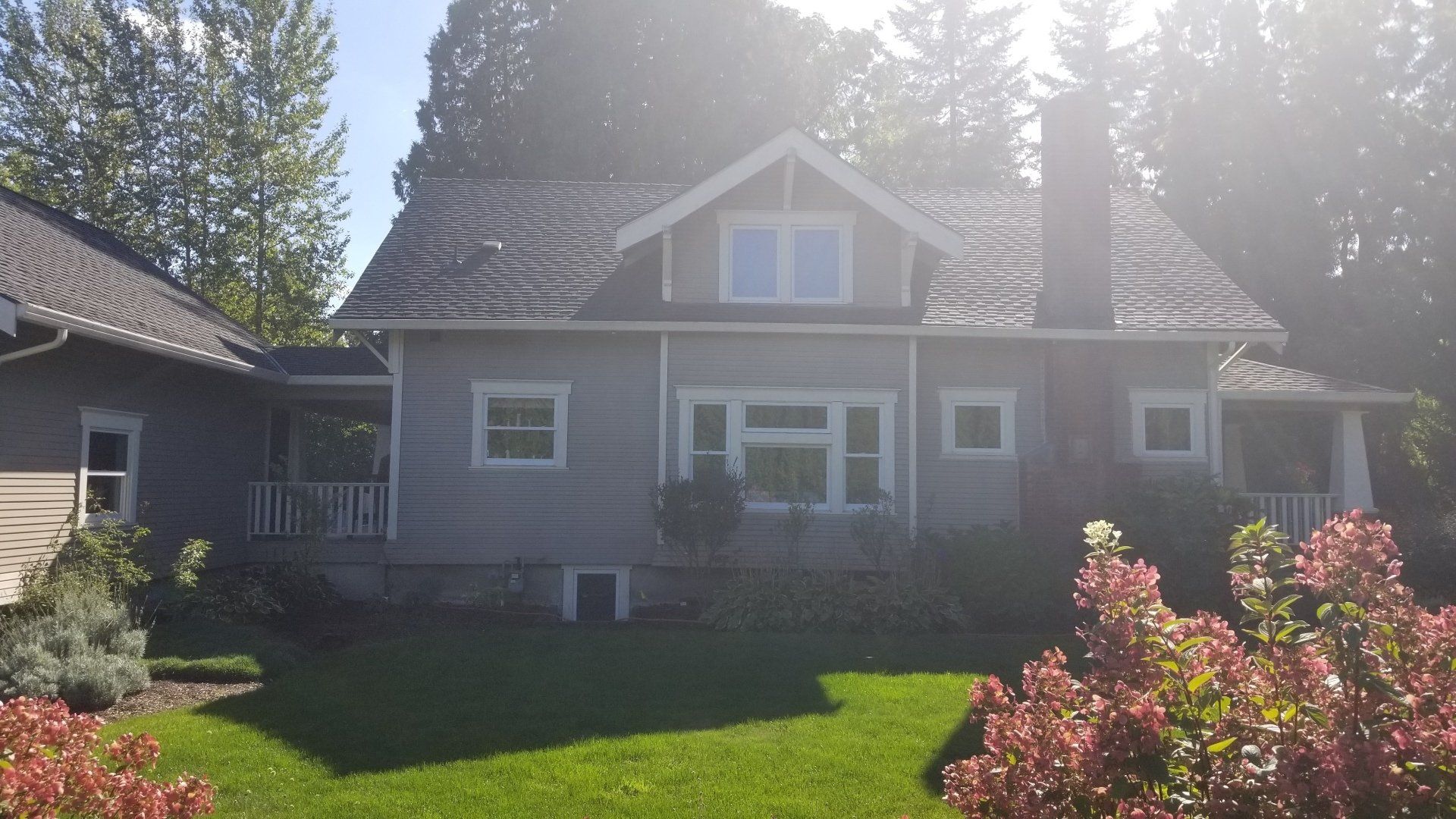 Gray house with black roof, chimney, and surrounding greenery under a bright sky.