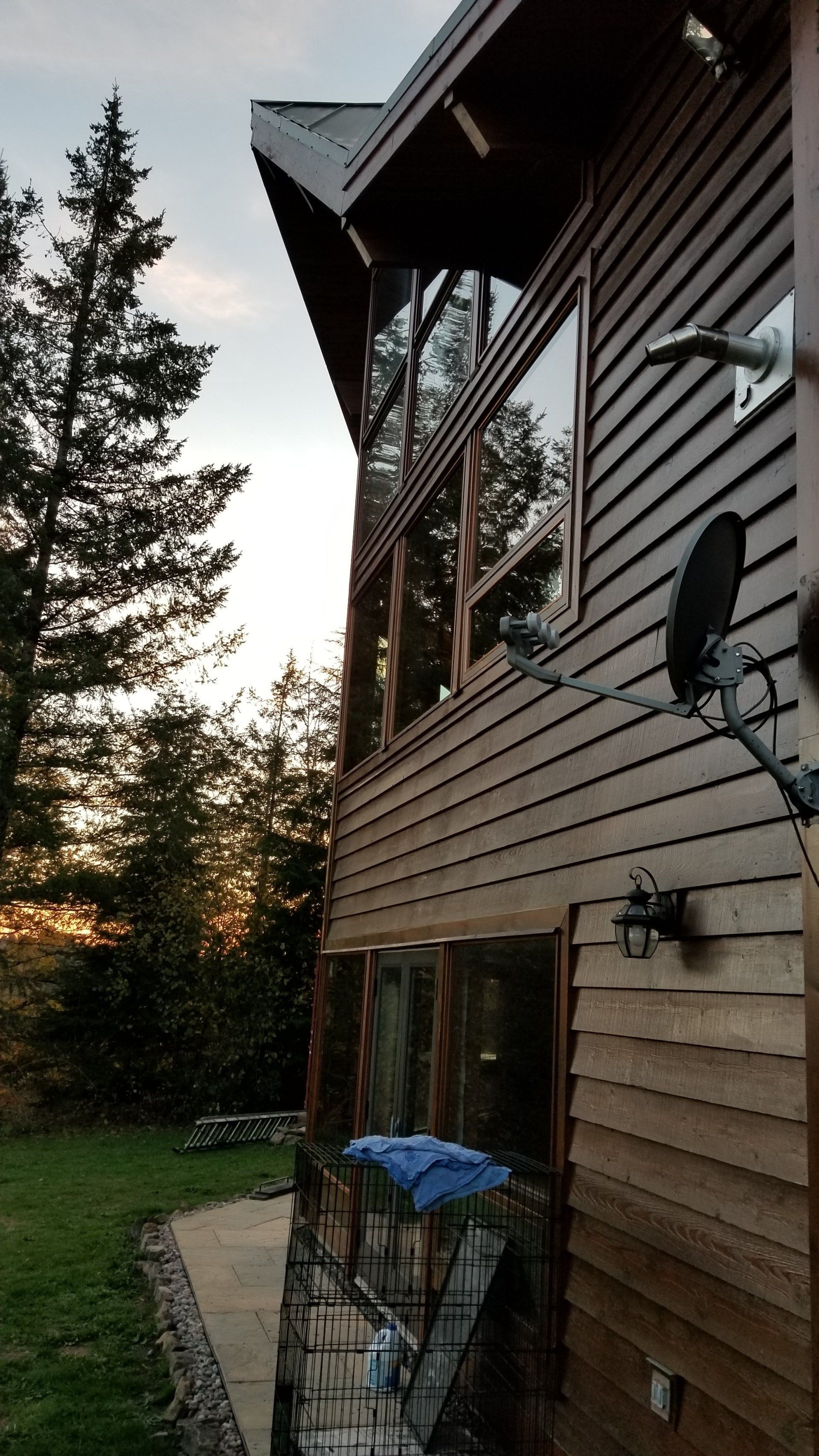 Wooden house exterior with large windows, satellite dish, and surrounding trees at dusk.