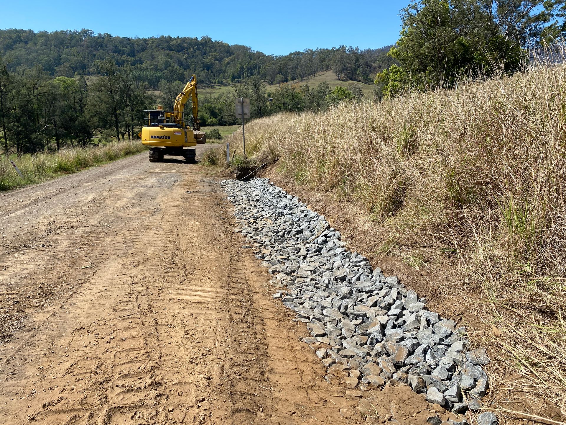 A Yellow Excavator Working on a Dirt Road, Beside a Row of Grey Rocks — PVM Contracting in Byron Bay, NSW