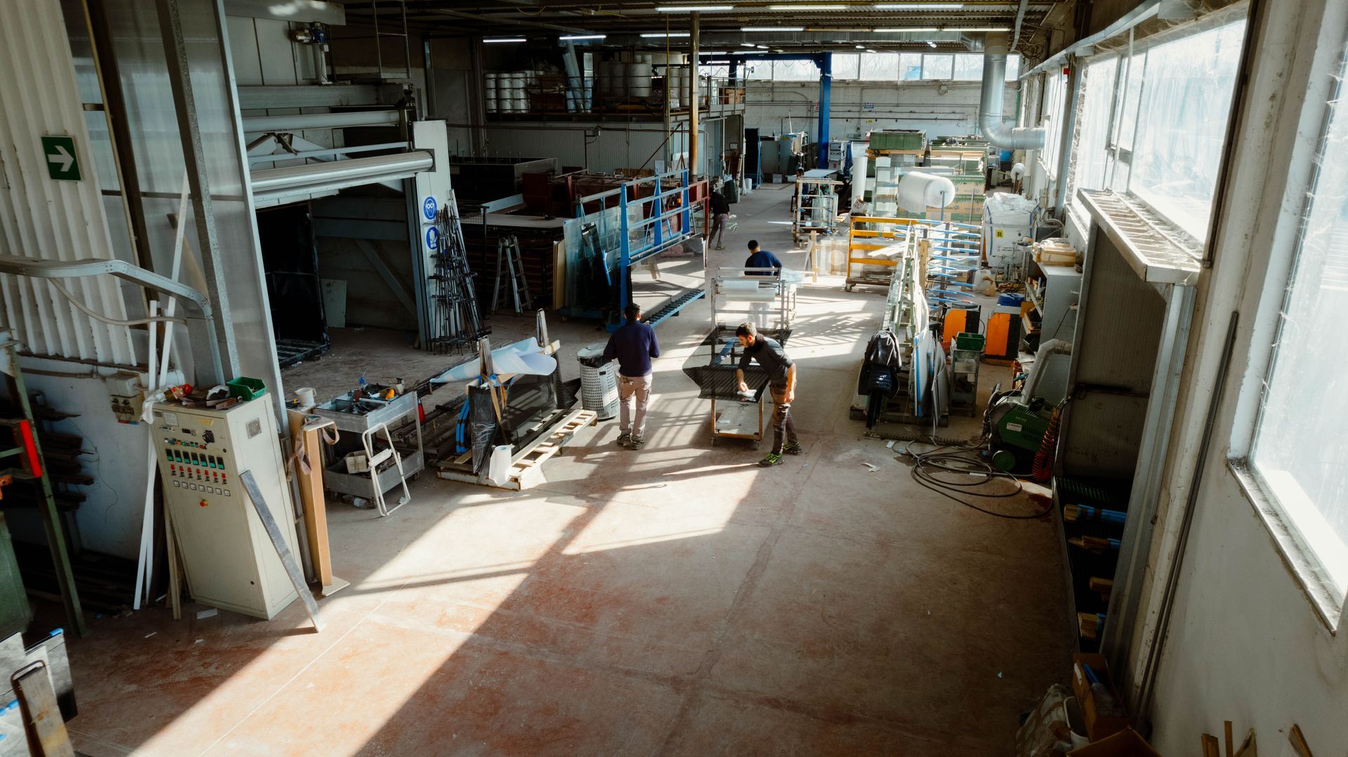 Una ripresa grandangolare e dall'alto di un'officina con operai vicino a vari macchinari e utensili sotto grandi finestre.