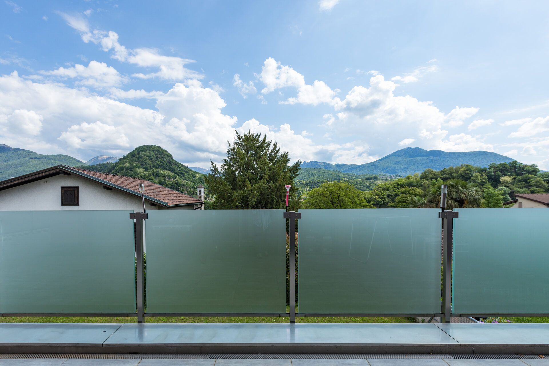 La ringhiera di un balcone in vetro satinato si affaccia su un paesaggio pittoresco di verdi colline, alberi e una casa sotto un cielo azzurro e nuvoloso.