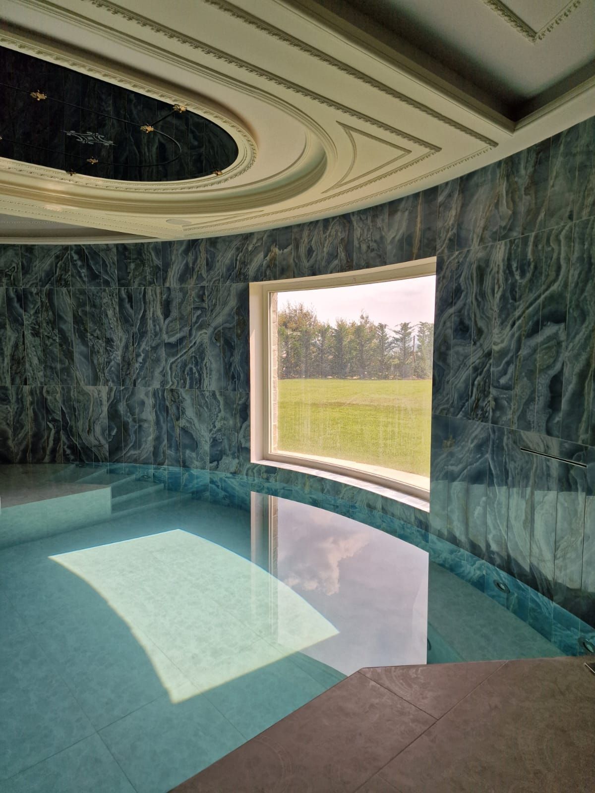 Piscina circolare interna con pareti in marmo blu, un'ampia finestra che si affaccia su un prato e un soffitto decorato e incassato.