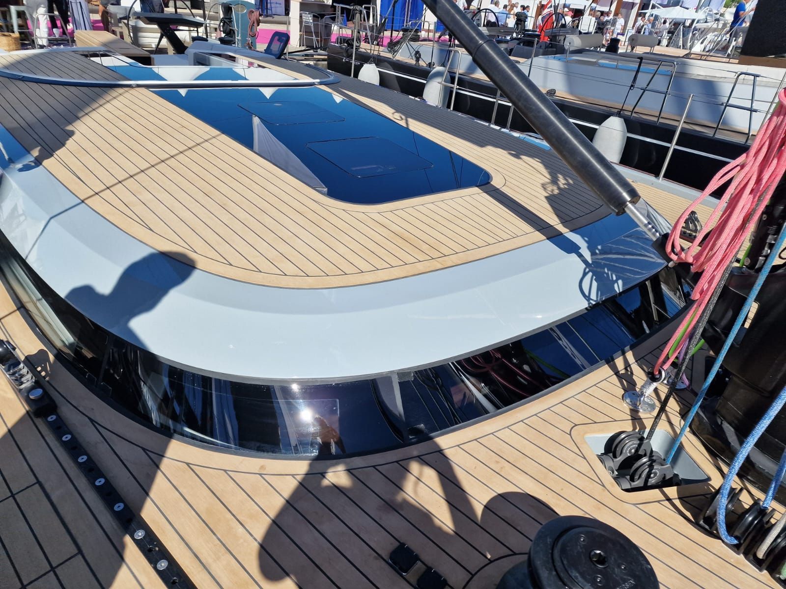 Un moderno yacht a motore bianco ormeggiato in un porto turistico con persone sul ponte sotto una tettoia, sullo sfondo di edifici costieri.