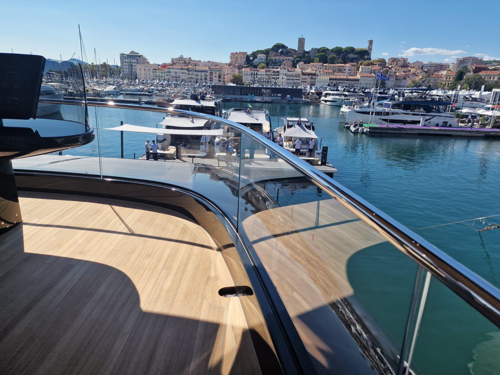 Una vista dal ponte di legno di uno yacht di lusso che si affaccia su un porto pieno di barche e su una città costiera sotto un cielo azzurro.