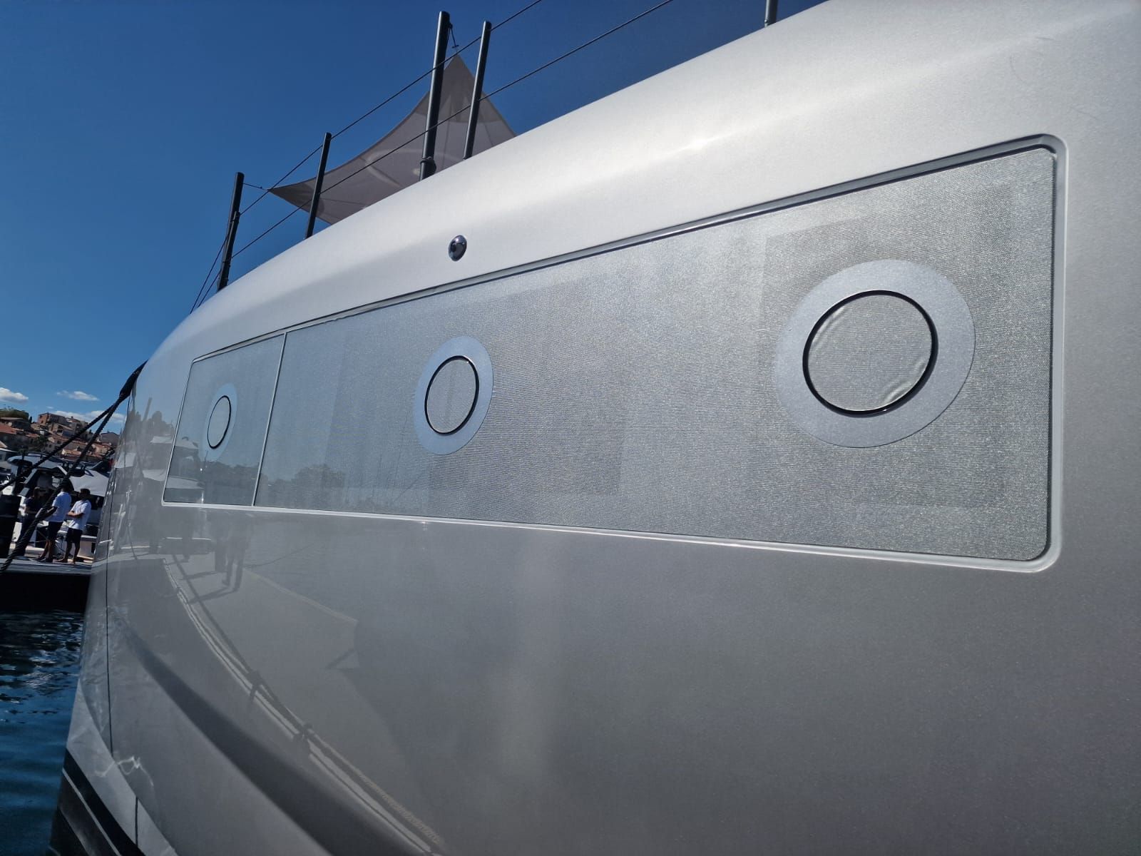 Primo piano dello scafo di uno yacht argentato caratterizzato da un pannello rettangolare con superficie ruvida e tre oblò circolari.