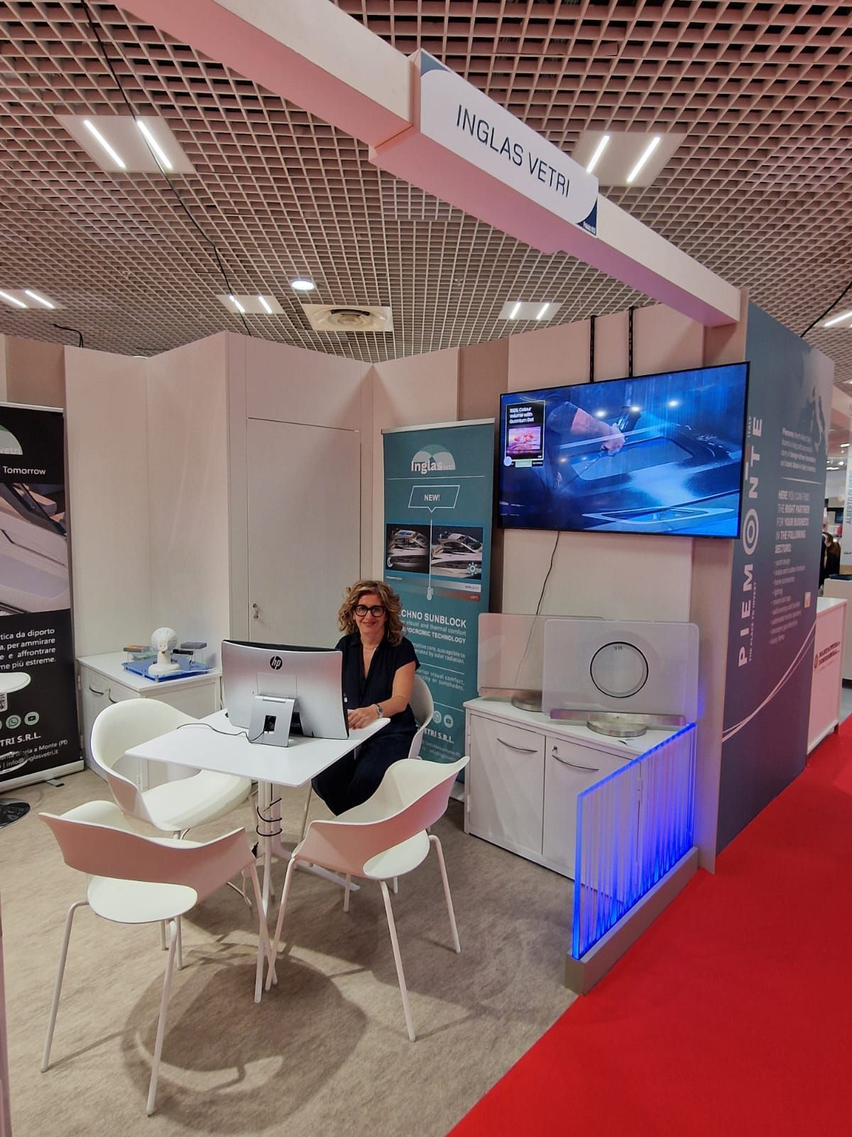 Una donna è seduta a un tavolo in uno stand fieristico, dotato di schermo televisivo, cartelli informativi sui prodotti e posti a sedere.