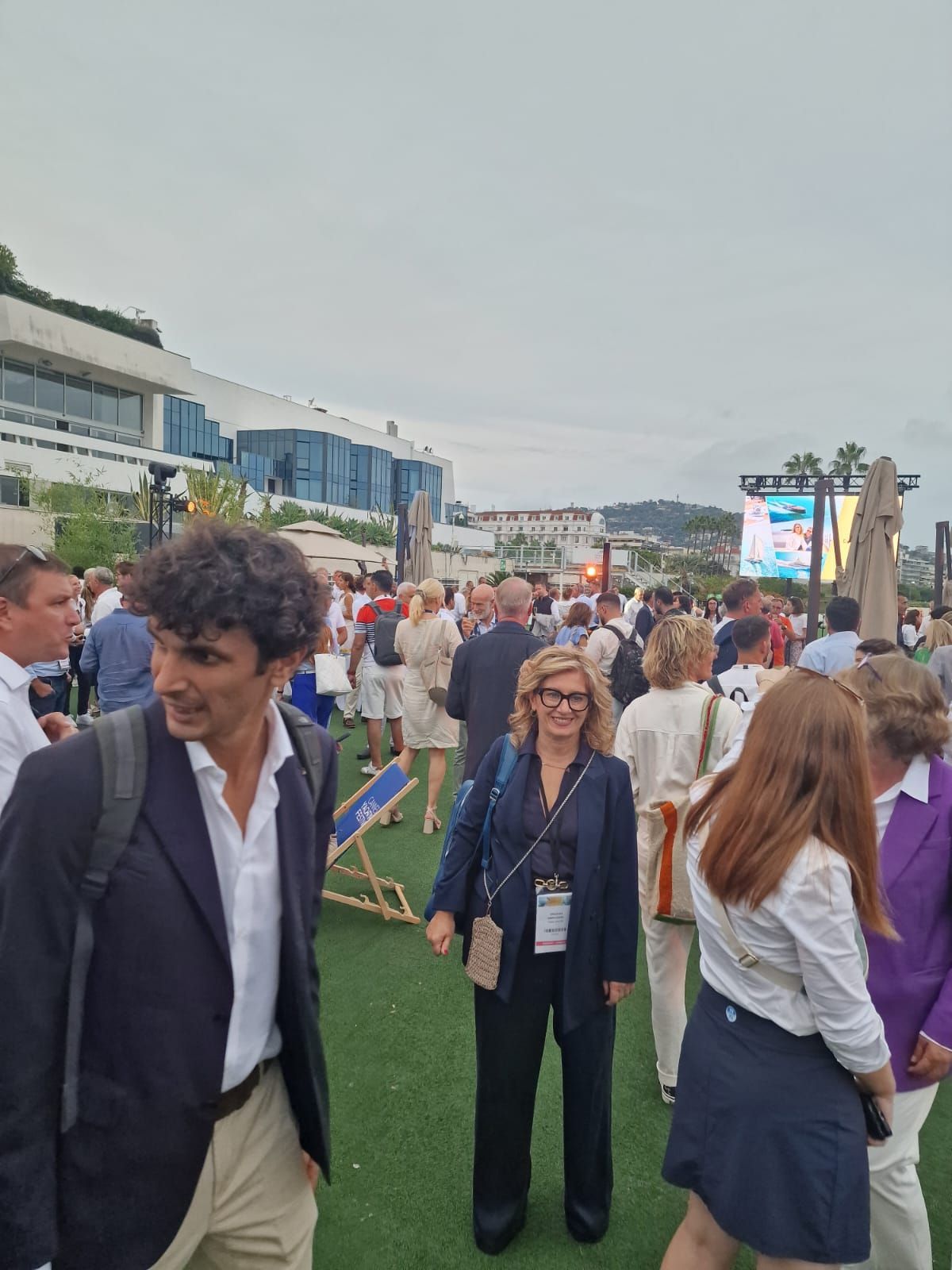 Durante un evento diurno, alcune persone si riuniscono su una terrazza verde all'aperto, con un edificio moderno e un grande schermo sullo sfondo.