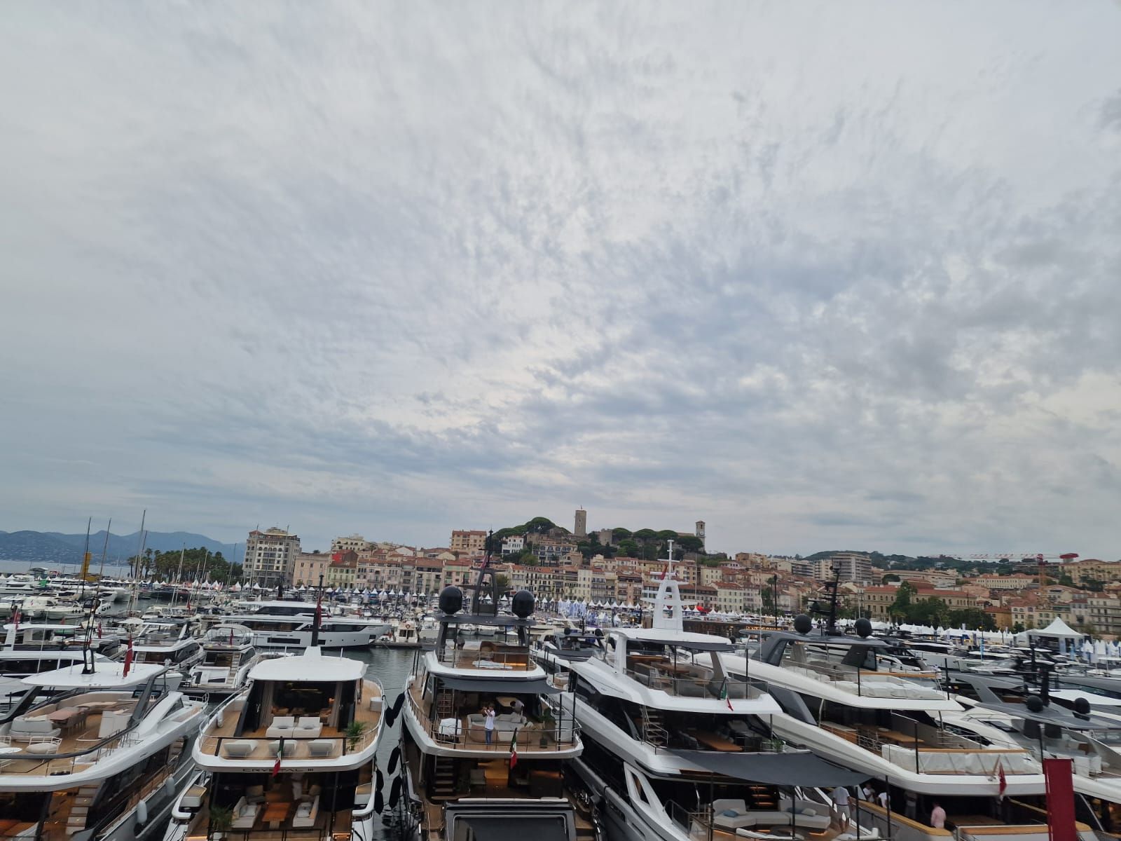 Un porticciolo pieno di yacht di lusso ormeggiati di fronte a una cittadina mediterranea adagiata su una collina, sotto un cielo nuvoloso.