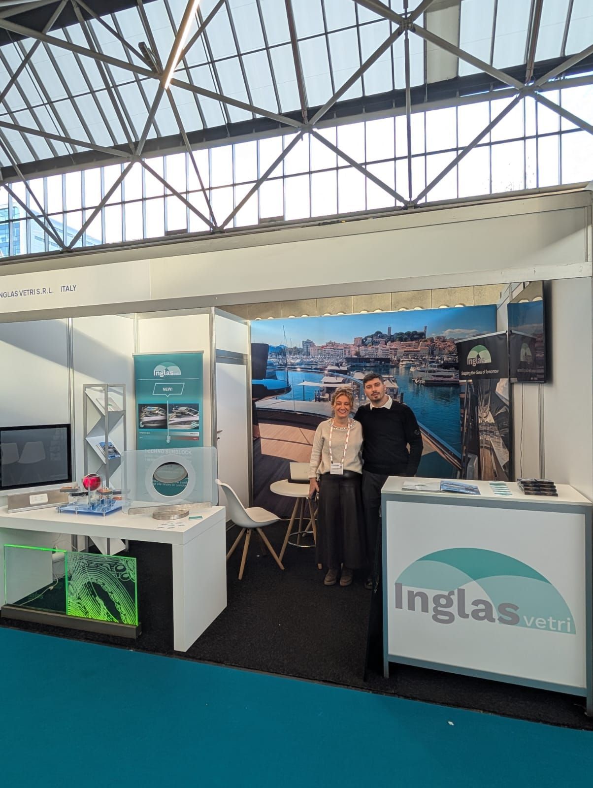 Un uomo e una donna si trovano presso lo stand fieristico di Ingla, con sullo sfondo un porto e l'esposizione di prodotti in vetro.
