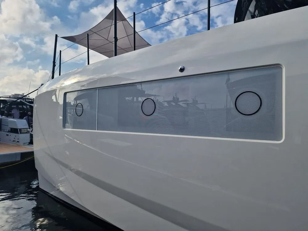 Una lunga finestra rettangolare con tre oblò circolari sul lato dello scafo bianco di uno yacht in un porto turistico.