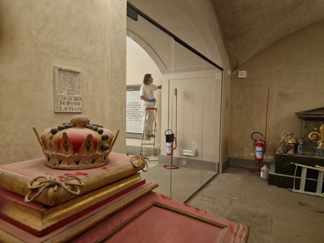 In primo piano, una corona poggia su una pila di libri, mentre all'interno di una stanza con le pareti in pietra una persona lavora su una scala a pioli.
