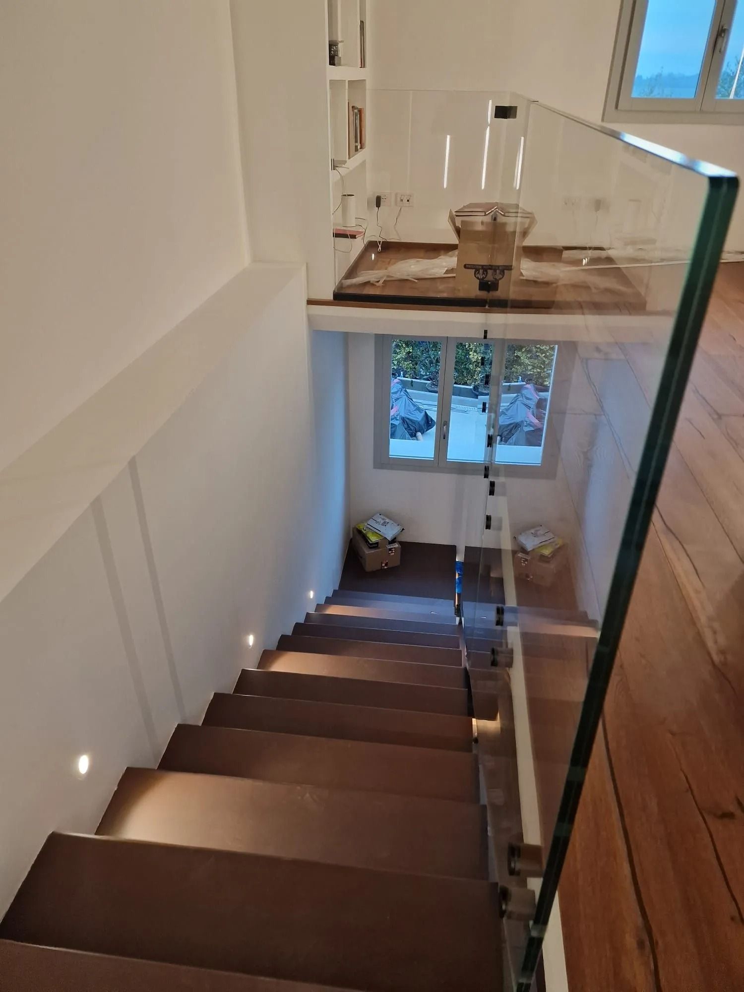 Una scala in legno con faretti a incasso a parete conduce a un piccolo pianerottolo con una finestra e una ringhiera in vetro.