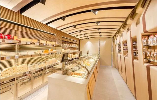Un negozio di formaggi luminoso e moderno, caratterizzato da scaffali in legno ordinatamente disposti e riforniti di vari formaggi, oltre a vetrine espositive.