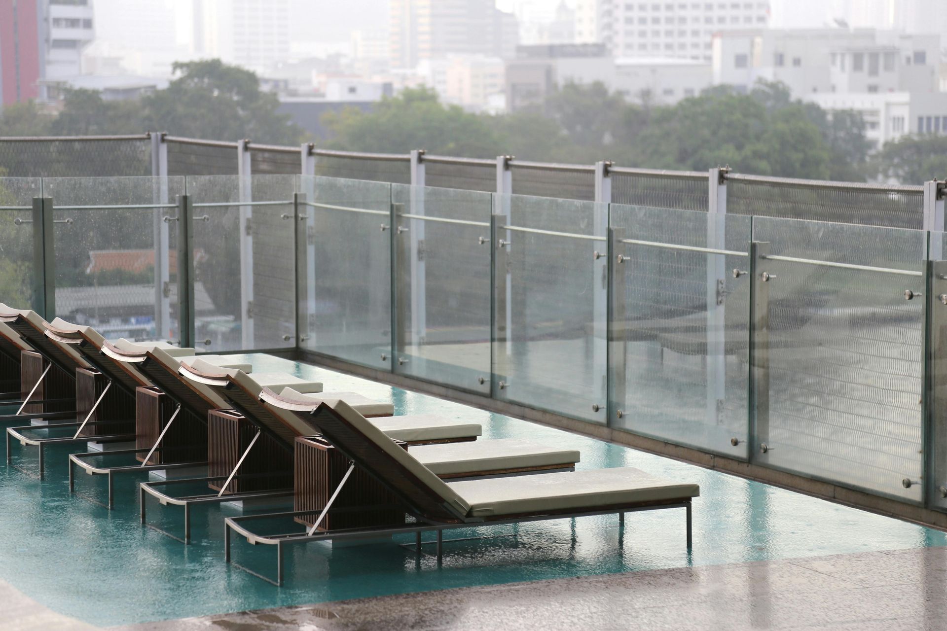 Sedie a sdraio vuote immerse in una piscina a sfioro sul tetto, sotto la pioggia, con una ringhiera in vetro che si affaccia sullo skyline della città.