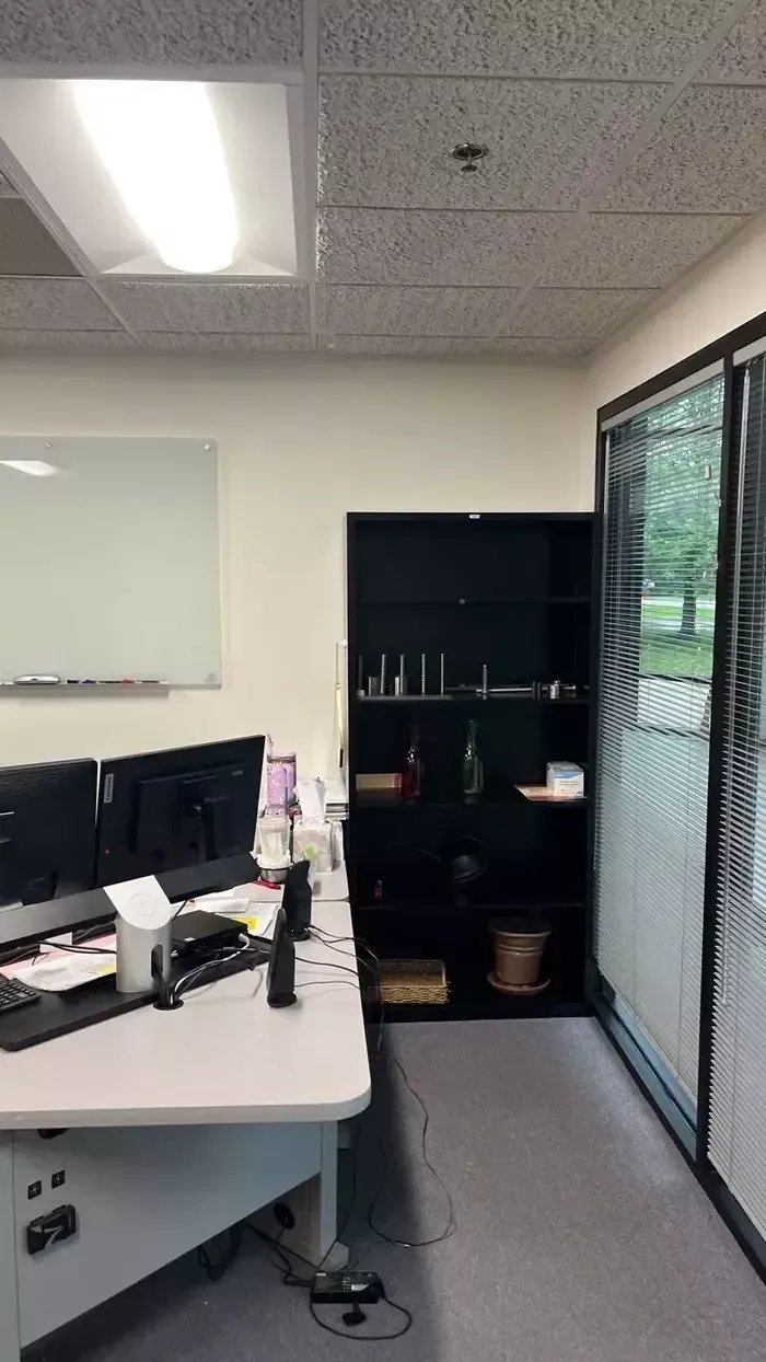 Small office with desk, computer monitors, whiteboard, cabinet, and window blinds.