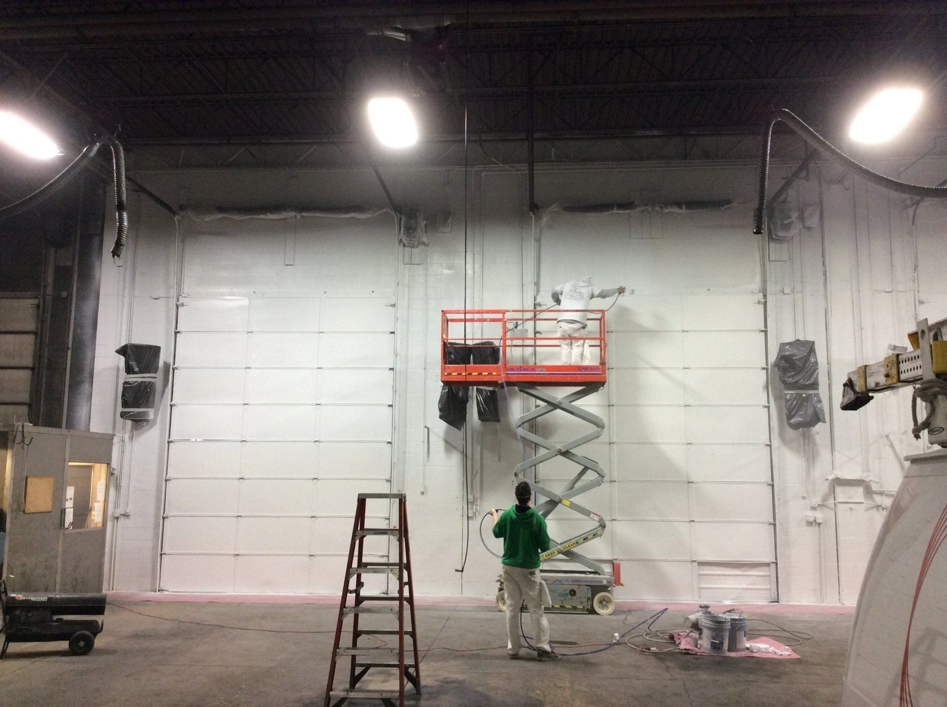 Worker on a scissor lift painting a large white warehouse wall under bright overhead lights.