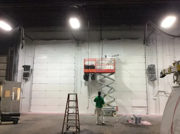 Worker on scissor lift painting a large white warehouse wall with a ladder nearby.