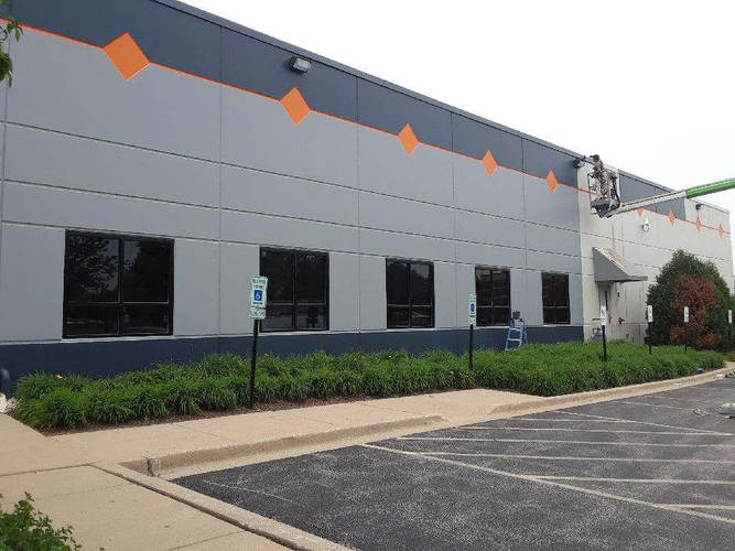 Gray industrial building with orange accents, windows, shrubs, and a parking lot in front